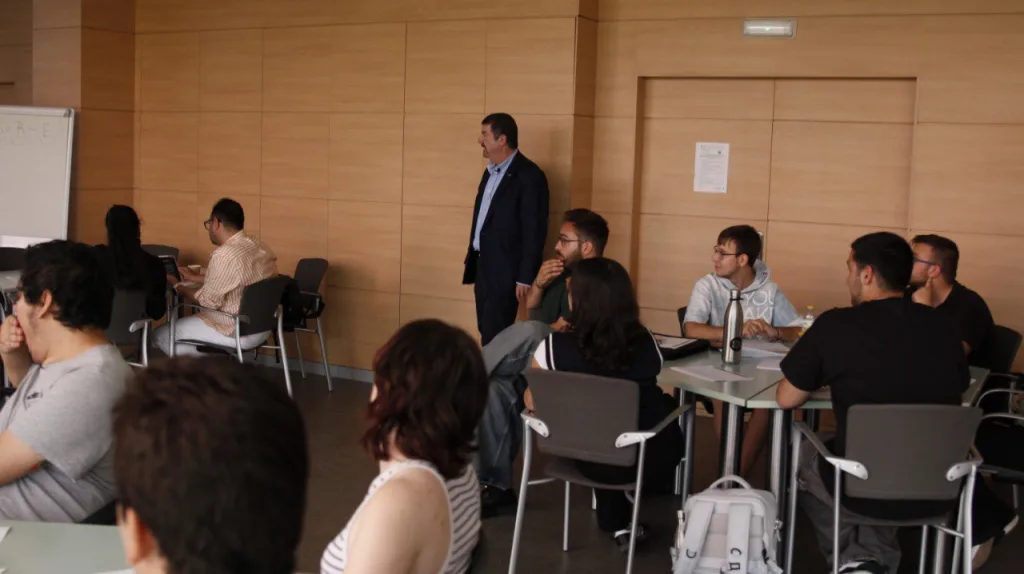 El ponente, de pie, se dirige a los estudiantes, sentados en grupos alrededor de las mesas