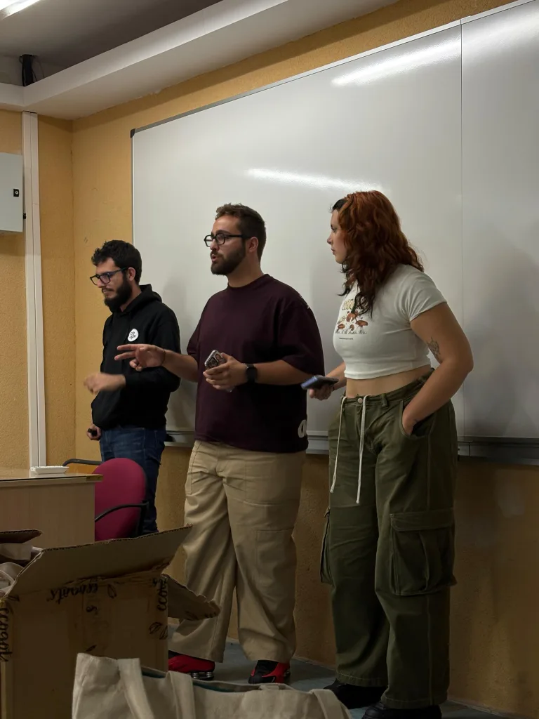 Tres miembros de la organización explican la actividad de pie, delante de la pizarra del aula