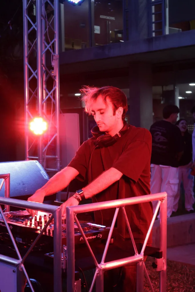 Un DJ pincha en una cabina de estructura metálica, con un edificio del campus al fondo