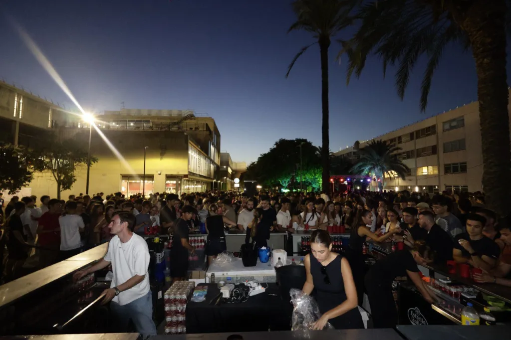 Cientos de estudiantes de noche junto a la barra de la CALUMFEST, entre palmeras y edificios del campus