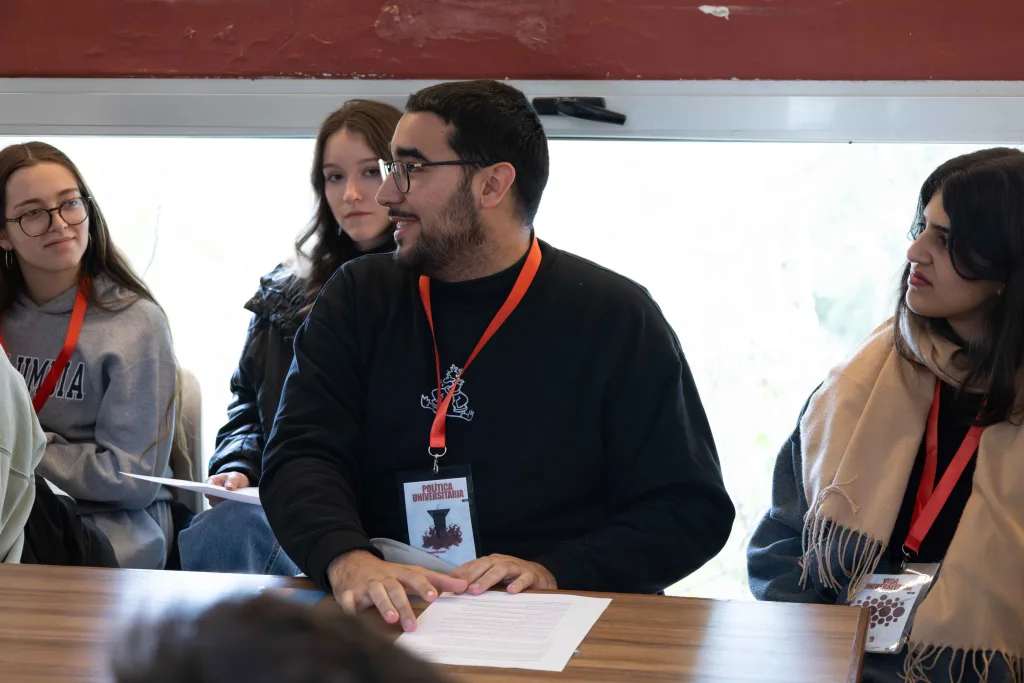 Participantes con acreditaciones sentados a una mesa durante una sesión de trabajo; uno de ellos interviene