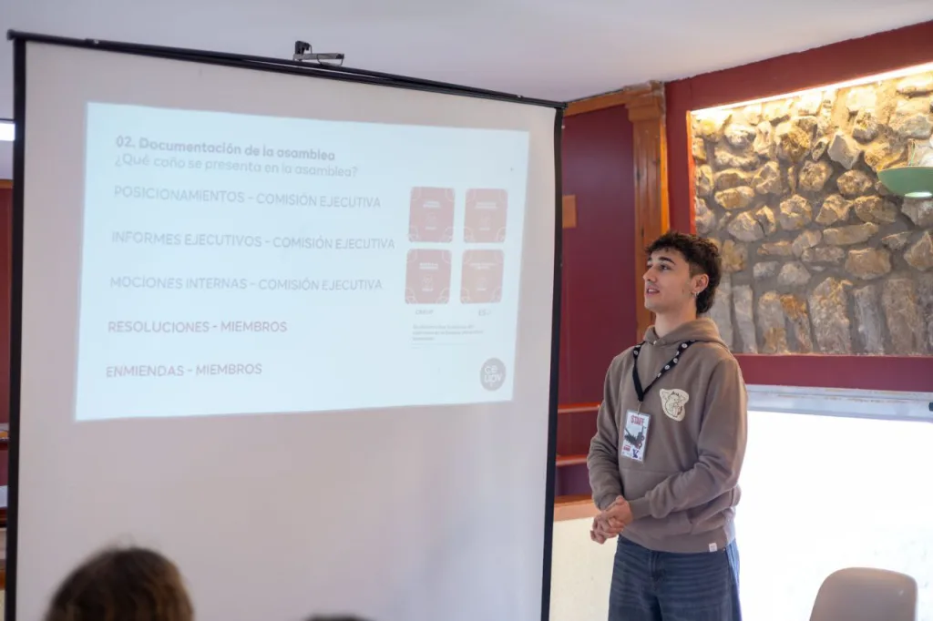 Un ponente presenta junto a una diapositiva sobre la documentación de la asamblea: posicionamientos, informes ejecutivos, mociones internas, resoluciones y enmiendas