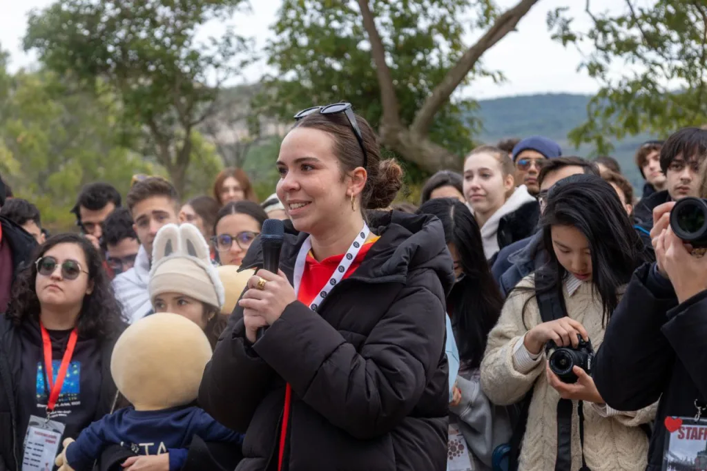 Una participante habla con micrófono rodeada de asistentes al aire libre, mientras otra persona hace fotos